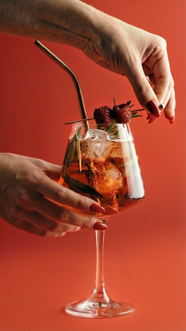 Une main dépose des framboises sur un cocktail, fond rouge.