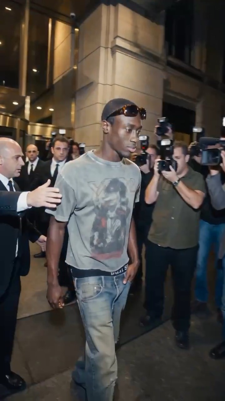 Un jeune homme en t-shirt vintage traverse une haie de photographes à la sortie d'un hôtel, la nuit.